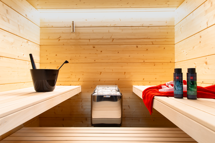 A modern Finnish sauna with light benches features a Harvia KIP45B Stainless Steel 4.5KW Sauna Heater with Built-in Control in the center, a black bucket and ladle on the left bench, and bottles beside a red towel on the right bench.