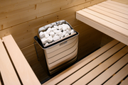 A Harvia KIP45B Stainless Steel 4.5KW Sauna Heater with Built-in Control sits between wooden benches in a light Finnish sauna, filled with white stones.