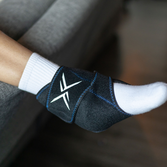 A person seated on a gray sofa, wearing the Icebound Essentials Frost Fit Hot/Cold Ankle Wrap over a white sock, demonstrates hot and cold therapy for the foot and ankle.