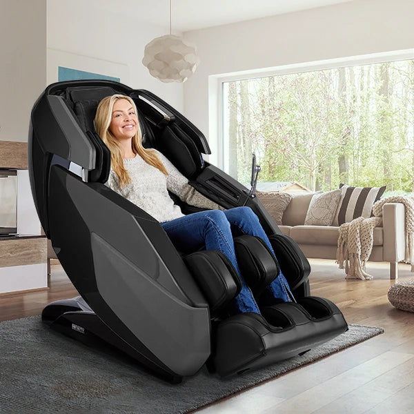 A woman smiles while sitting in the Infinity Imperial Syner-D Massage Chair by Infinity in a bright, modern living room with a sofa, rug, and large windows revealing trees outside.