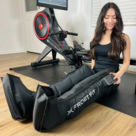 A woman in workout clothes smiles while sitting on the floor with both legs inside Icebound Essentials Recovery Wraps™, Wireless Compression Boots; an exercise bike and flat-screen monitor are seen in the background.