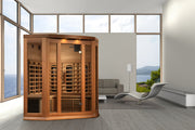 The Golden Designs Maxxus 3 Person Full Spectrum IR Sauna (MX-M356-01-FS CED) sits in a spacious, minimalist living room with large windows framing ocean and mountain views, alongside a gray sofa, sleek lounge chair, and white floor lamp.