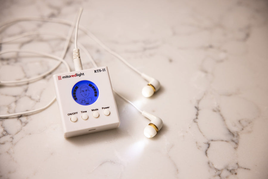A compact white MitoBOOST™ Intranasal Device System by Mito Red Light, featuring a blue digital screen and earbud-style attachments, rests on marble—designed for sinus health, immune support, and brain health.