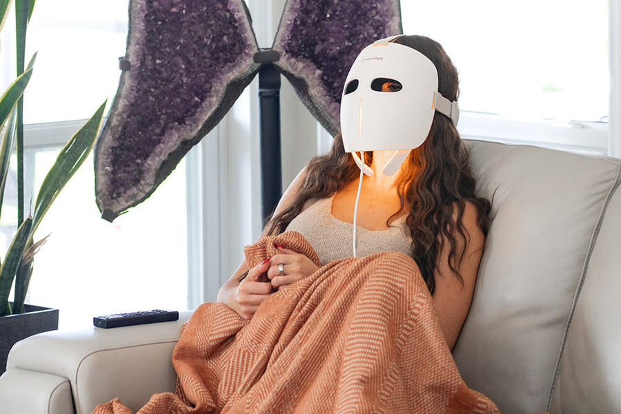 Relaxing on a sofa with a blanket, a person wears the MitoGLOW™ LED Red Light Therapy Mask by Mito Red Light. A large purple crystal display and tall green plant sit behind them, while a TV remote rests on the armrest.