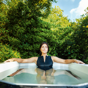 A woman in a black swimsuit enjoys an invigorating cold plunge in The Resolute Pro by Icebound Essentials, relaxing with arms outstretched in an outdoor tub surrounded by lush greenery beneath a bright blue sky.