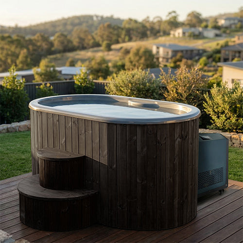 The SaunaLife Model S2BC – 2-Person Immersion Tub with Black Chiller & Heater is a round, wooden hot and cold tub on a deck amid lush greenery—an ideal home spa by SaunaLife for relaxing hydrotherapy with views of hills and houses.