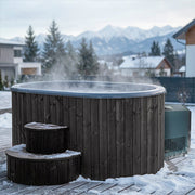 The SaunaLife Model S2BC – 2-Person Immersion Tub with Black Chiller & Heater sits steaming on a snowy deck outdoors, surrounded by homes, evergreens, and snow-capped mountains in the background.