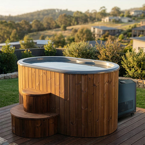 The SaunaLife Model S2NC 2-Person Immersion Tub with Natural Finish Chiller & Heater sits on a backyard deck, surrounded by greenery and overlooking distant houses and hills under a clear sky.