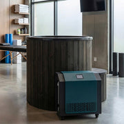 The SaunaLife Model S1BC is a modern indoor home spa featuring a 1-person immersion tub with black chiller and heater, stacked towels, fitness equipment on shelves, and large windows that fill the space with natural light.