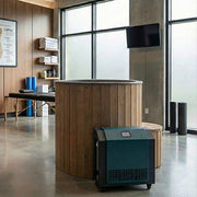 A modern home spa room features the SaunaLife Model S1NC— a 1-person immersion tub with natural finish chiller & heater— beside shelves with towels and supplies, a massage table, foam rollers, and large frosted windows letting in natural light.