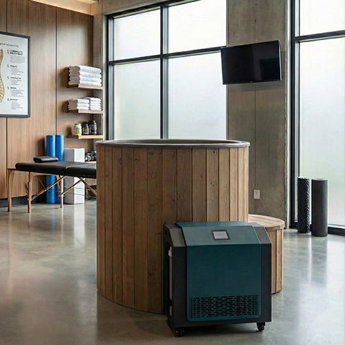 A modern home spa room features the SaunaLife Model S1NC— a 1-person immersion tub with natural finish chiller & heater— beside shelves with towels and supplies, a massage table, foam rollers, and large frosted windows letting in natural light.