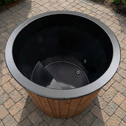 The SaunaLife Model S1NC – 1-Person Immersion Tub with Natural Finish Chiller & Heater features a round, black design with wood paneling, bench seat, and is ideal for hot or cold hydrotherapy on your home’s gray stone patio.