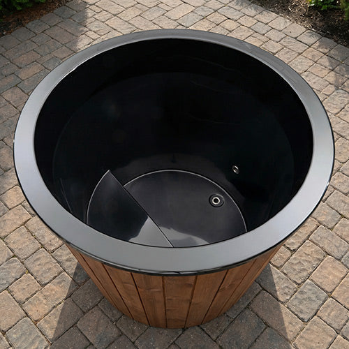 The SaunaLife Model S1NC – 1-Person Immersion Tub with Natural Finish Chiller & Heater features a round, black design with wood paneling, bench seat, and is ideal for hot or cold hydrotherapy on your home’s gray stone patio.