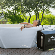 A man relaxes in a white portable ice bath labeled The Endurance Plunge + Chiller by Icebound Essentials on a wooden deck, surrounded by trees and natural scenery.