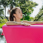 A woman with closed eyes relaxes in The Pink Endurance Plunge Tub by Icebound Essentials, surrounded by greenery and sunlight. Her arm tattoo adds flair to this peaceful outdoor wellness retreat.