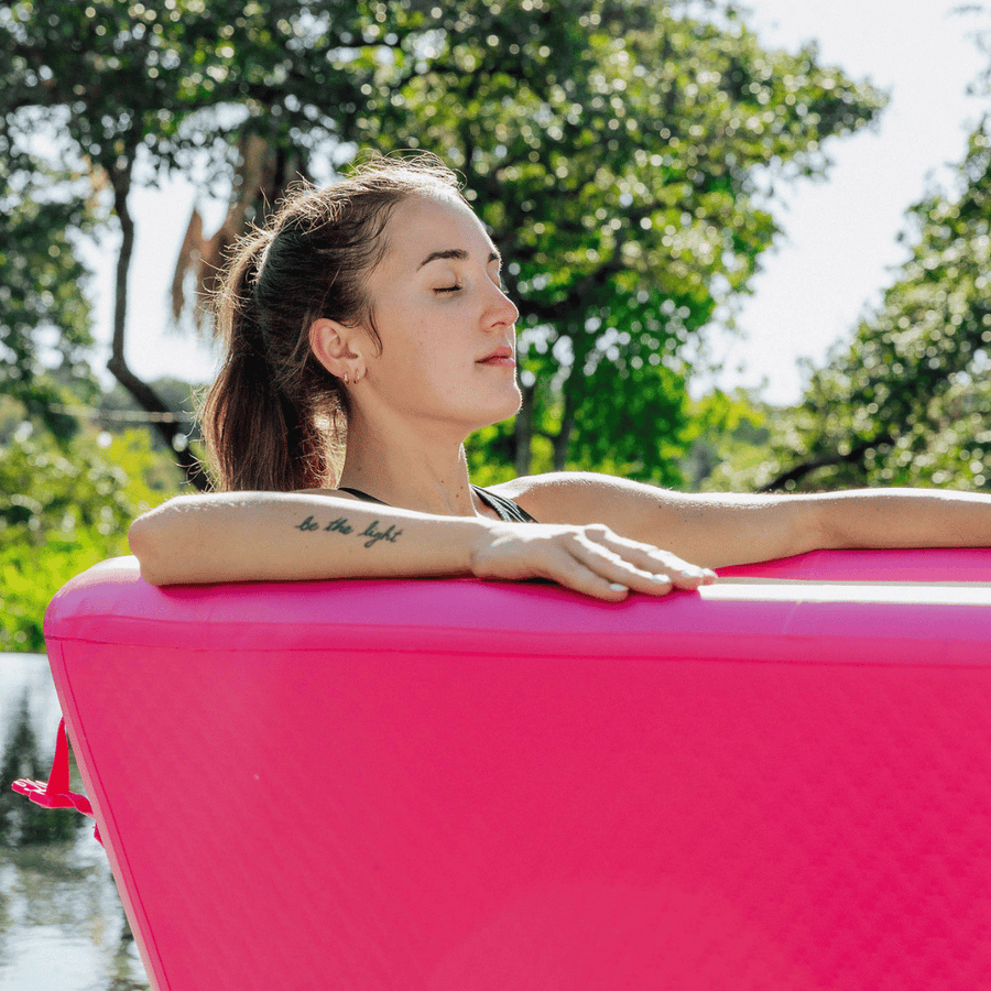 A woman with closed eyes relaxes in The Pink Endurance Plunge Tub by Icebound Essentials, surrounded by greenery and sunlight. Her arm tattoo adds flair to this peaceful outdoor wellness retreat.