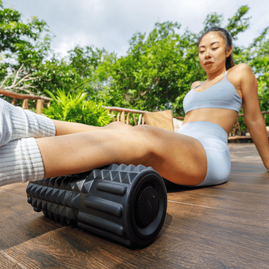 A woman in athletic wear uses the Icebound Essentials VibePro Flow deep massage foam roller on her calves while sitting on a wooden deck outdoors, surrounded by greenery.