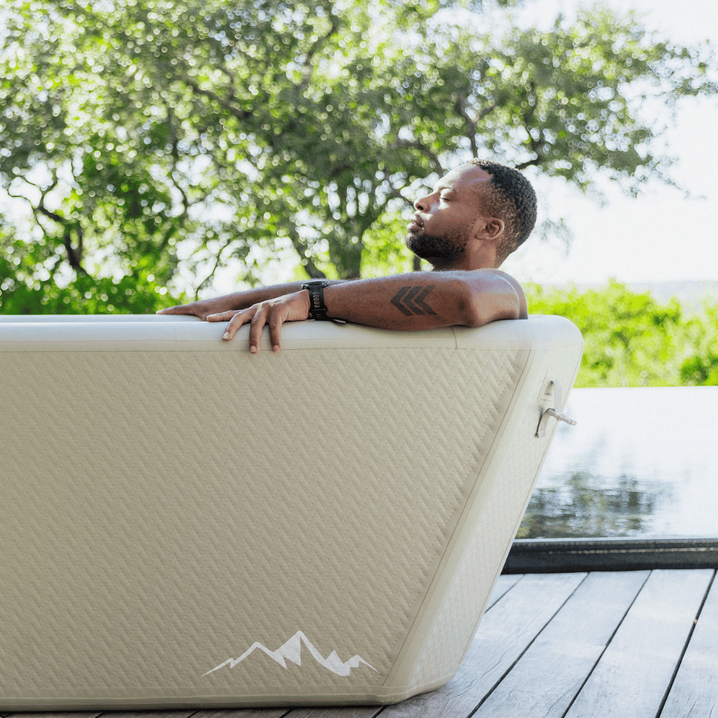 A man relaxes in The Endurance Plunge Tub by Icebound Essentials, eyes closed and surrounded by trees with a pond or pool nearby, enjoying a peaceful moment immersed in nature.