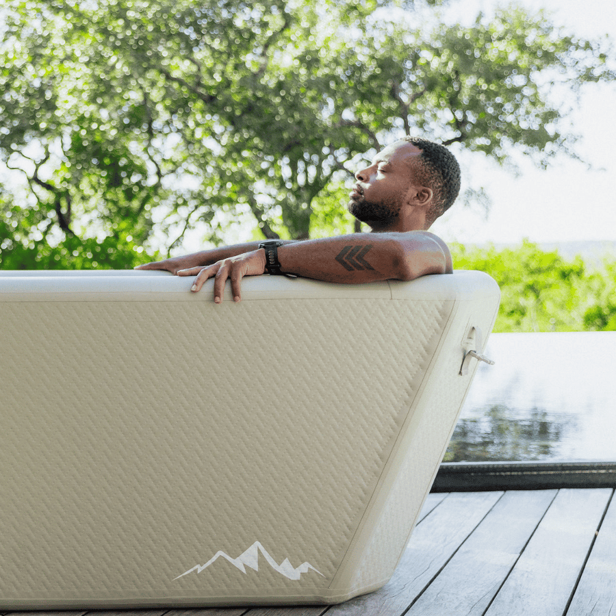 A man relaxes in The Endurance Plunge Tub by Icebound Essentials, eyes closed and surrounded by trees with a pond or pool nearby, enjoying a peaceful moment immersed in nature.
