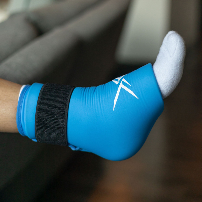 A person wearing the Icebound Essentials Frost Fit Ankle Ice Sleeve—a blue foot pad with a black strap and white logo—rests indoors. This sleeve is ideal for ankle pain relief or plantar fasciitis.