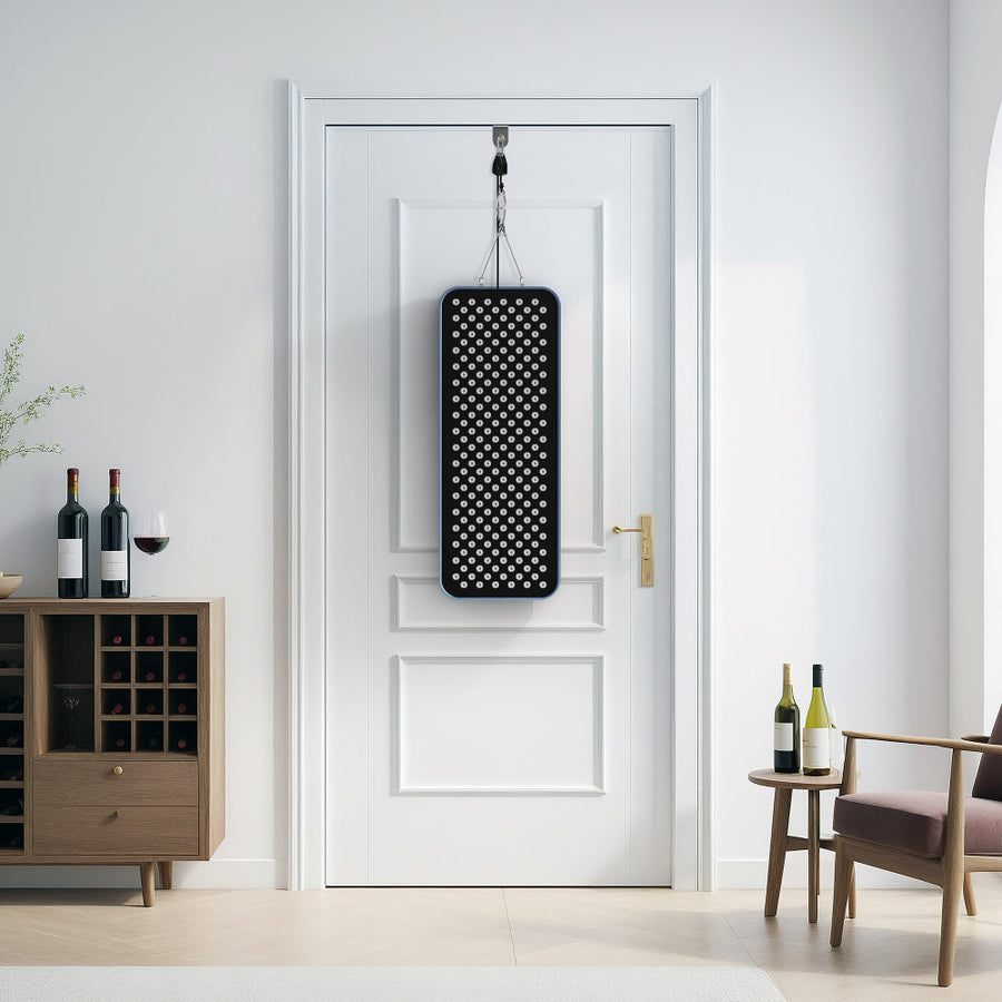 The RLT Home Total Spectrum COMPACT Red Light Therapy Panel by RLT Home hangs on a white door in a modern room with a wine rack, bottles, a plant, small table, and chair. Natural light streams through an arched window.