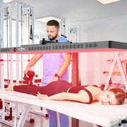 A woman lies face down on a massage table under the RLT Home Total Spectrum ELITE Red Light Therapy Panel while a man in medical scrubs uses a massage gun on her calf in a clinic setting.