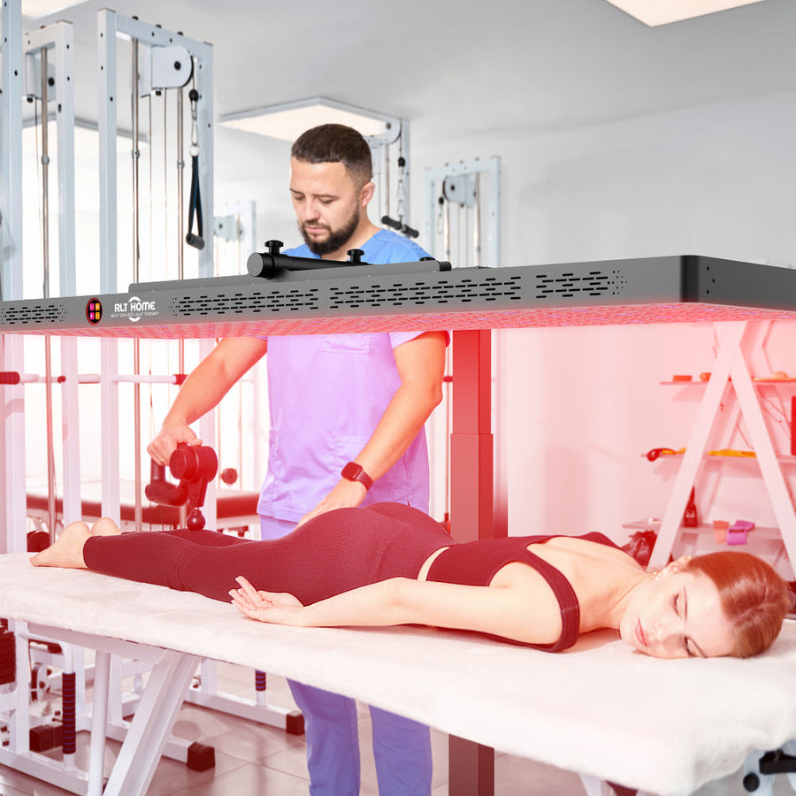 A woman lies face down on a massage table under the RLT Home Total Spectrum ELITE Red Light Therapy Panel while a man in medical scrubs uses a massage gun on her calf in a clinic setting.