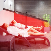 Two people in white robes relax on lounge chairs at a modern spa, enjoying the RLT Home Total Spectrum ELITE Red Light Therapy Panel by RLT Home, with dark tiled walls and a potted plant in the background.