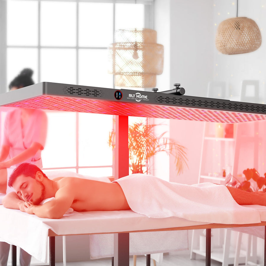 A man enjoys a full-body session under the RLT Home Total Spectrum ELITE Red Light Therapy Panel by RLT Home, while a person in pink uniform stands in the bright, modern room with plants and soft lighting.