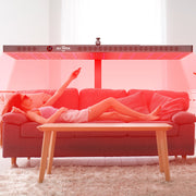 A woman enjoys a full-body treatment on a red sofa using the RLT Home Total Spectrum ELITE Red Light Therapy Panel with a remote. The bright room features white curtains and a wooden coffee table in front of the sofa.