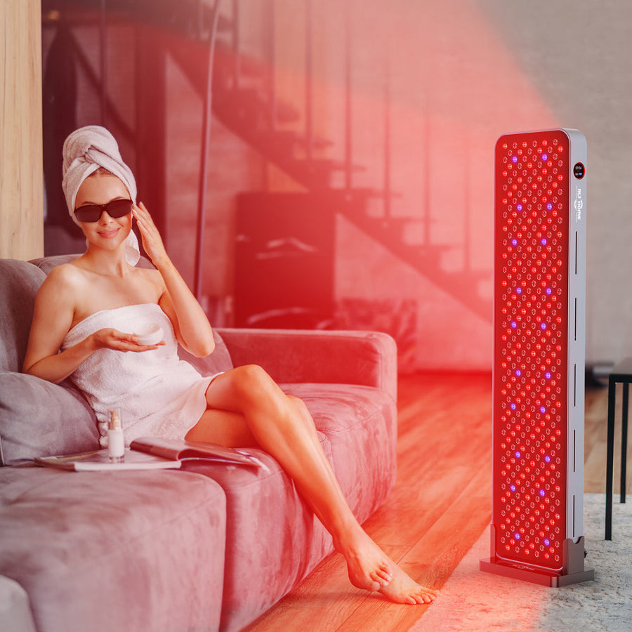 A woman in a towel and sunglasses sits on a sofa, using the RLT Home Total Spectrum MAX Red Light Therapy Panel as it emits red light for skin restoration—showcasing RLT Home’s wellness device in a modern living room.