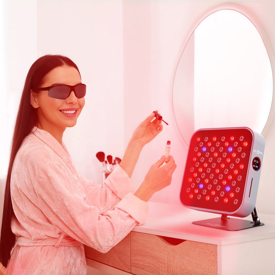 A woman in a pink robe and sunglasses applies serum at her vanity while using the RLT Home Total Spectrum MINI Red Light Therapy Panel for targeted treatment, surrounded by a mirror and makeup items that showcase her home wellness routine.