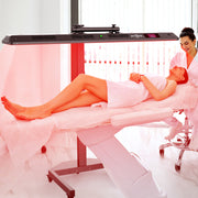 A woman relaxes on a treatment bed under the RLT Home Total Spectrum ULTRA Red Light Therapy Panel as a smiling technician in a white coat assists her, highlighting the device's rejuvenating benefits in a bright, modern clinic.