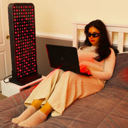 A woman wearing red-tinted glasses uses a laptop on her bed, illuminated by the RLT Home Total Spectrum COMPACT Red Light Therapy Panel from RLT Home, filling the room with a soothing red glow for holistic health benefits.