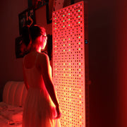 A woman in a white dress stands before the RLT Home Total Spectrum ELITE (7 Wave 864 LEDs), its red and green lights illuminating her in a dim room, highlighting the benefits of full-body red light therapy.