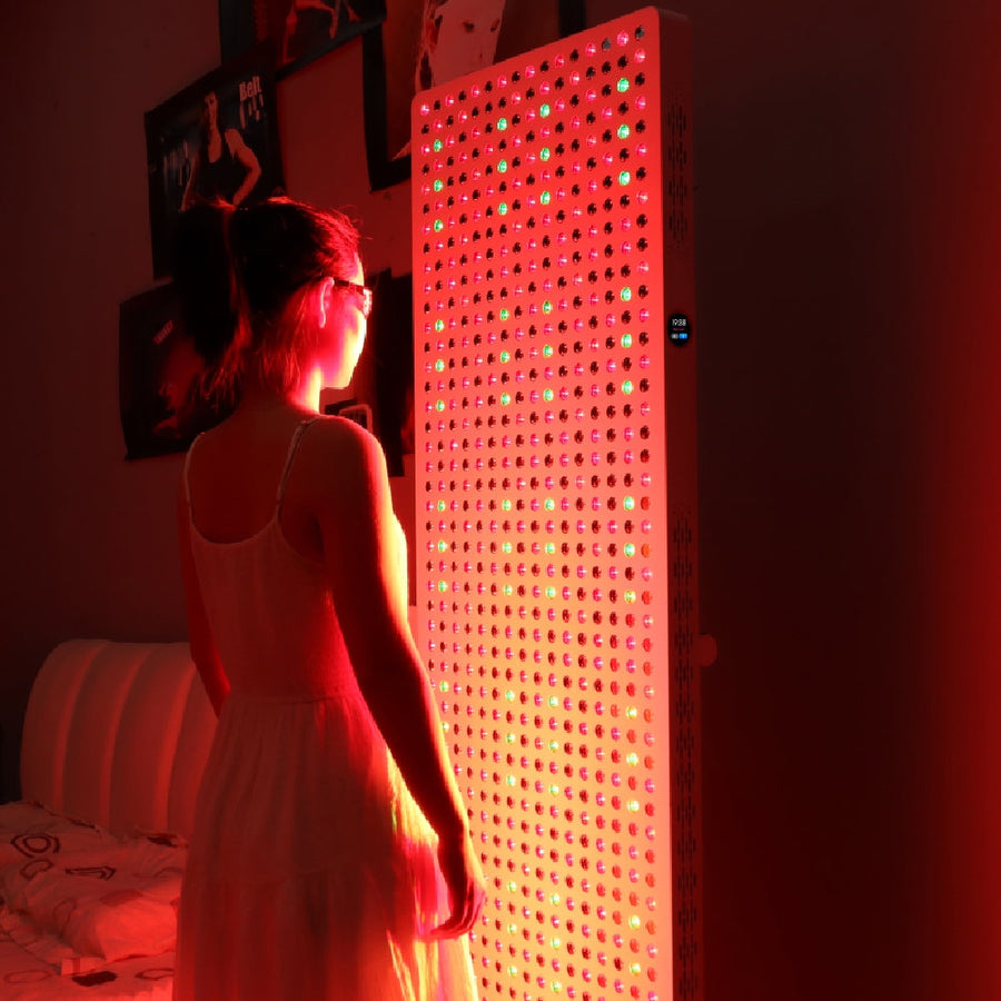 A woman in a white dress stands before the RLT Home Total Spectrum ELITE (7 Wave 864 LEDs), its red and green lights illuminating her in a dim room, highlighting the benefits of full-body red light therapy.