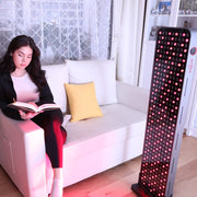 A woman reads on a white sofa, bathed in light from the RLT Home Total Spectrum MAX (7 Wave 360 LEDs) red light therapy device that promotes wellness. The room includes sheer curtains, a yellow cushion, and shelves in the background.