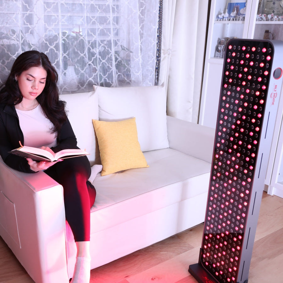 A woman relaxes on a white sofa reading, bathed in the glow of the RLT Home Total Spectrum MAX Red Light Therapy Panel—showcasing how RLT Home's innovative device brings holistic wellness benefits into her daily life.