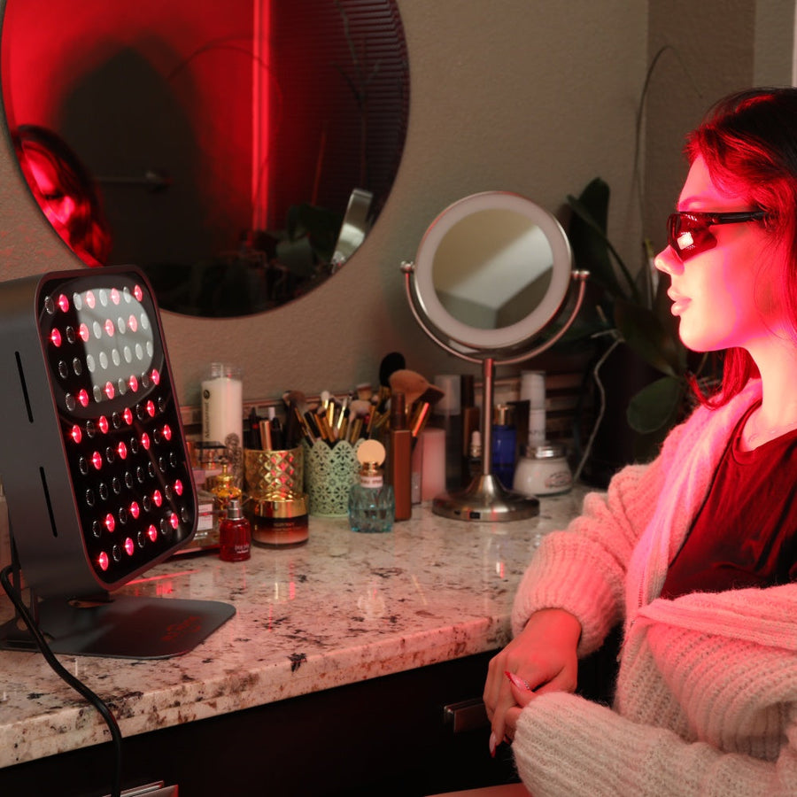 A woman wearing protective glasses sits at a vanity, using the RLT Home Total Spectrum MINI (7 Wave 72 LEDs) red light therapy device on her face. The countertop holds beauty products, brushes, and mirrors.