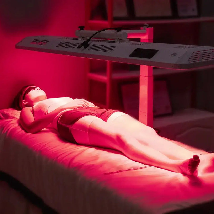 A person relaxes on a treatment bed beneath RLT Home's Total Spectrum ULTRA (7 Wave 480 LEDs), wearing protective goggles in a softly lit room for a red light therapy session promoting full-body wellness.
