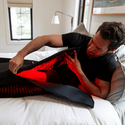 A man lies on a bed and wraps the HEALiX Infrared Elevate Light Pod, a black device with red LED lights, around his lower body for red light therapy and relaxation.
