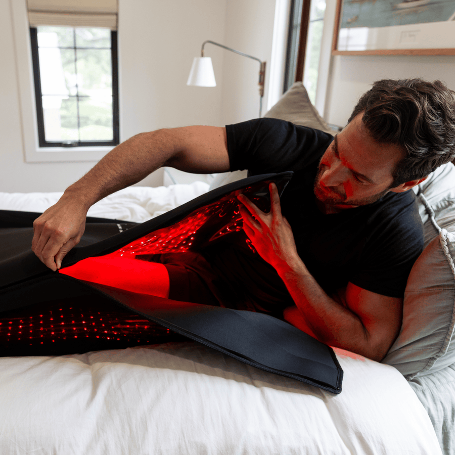 A man lies on a bed and wraps the HEALiX Infrared Elevate Light Pod, a black device with red LED lights, around his lower body for red light therapy and relaxation.
