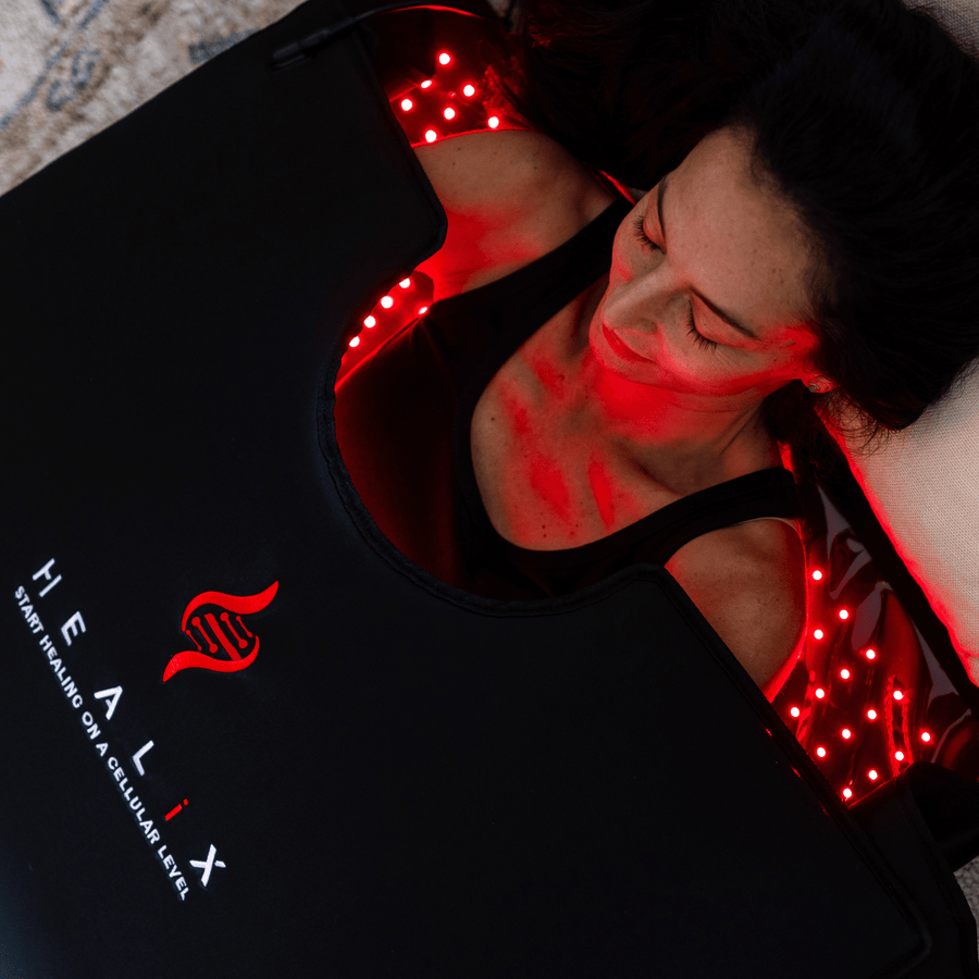 A woman lies on a black HEALiX Infrared Elevate Light Pod with red LED lights for red light therapy and pain relief. The mat has a red logo and the phrase "STARTS HEALING ON A CELLULAR LEVEL.