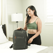 A woman with long dark hair, in a green crop top and black shorts, stands by a bed smiling while holding the handle of a black HEALiX Infrared backpack—ideal for carrying HEALiX Flow Pro Compression Boots—in a bright, modern bedroom.