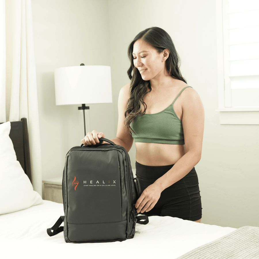 A woman with long dark hair, in a green crop top and black shorts, stands by a bed smiling while holding the handle of a black HEALiX Infrared backpack—ideal for carrying HEALiX Flow Pro Compression Boots—in a bright, modern bedroom.
