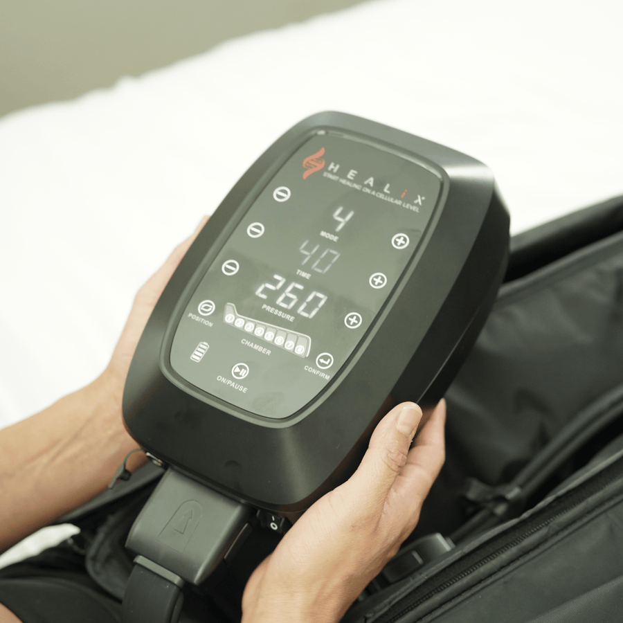 A person holds a black HEALiX Infrared HEALiX Flow Pro Compression Boot with a digital display and buttons, likely used for recovery or pneumatic therapy, on a white surface.