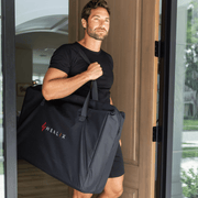 A man in black stands in a doorway, carrying a large black HEALiX Infrared bag—ideal for transporting the Elevate Light Pod. Bright natural light and greenery are visible outside.