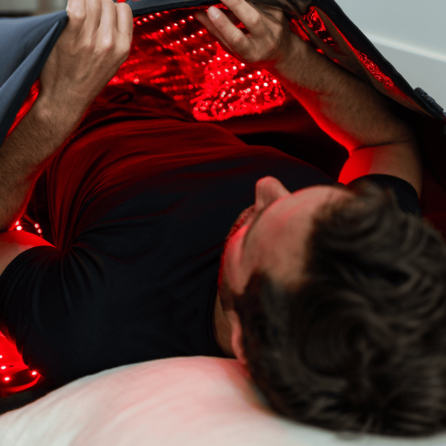 A man relaxes under the HEALiX Infrared Elevate Light Pod, with red LED light enveloping his upper body for pain relief, while he rests his head on a white pillow.