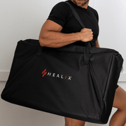 A person in black clothing carries a large black bag with the red and white HEALiX Infrared logo, designed for easy transport of the Elevate Light Pod—perfect for convenient, on-the-go red light therapy pain relief.