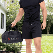 A man in a black t-shirt and shorts stands outside on a sunny day, holding the HEALiX Infrared HEALiX Flow gym bag with the red and white logo and DNA motif. Lush green grass and trees can be seen behind him.